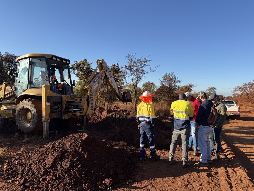 A backhoe loader digging a sampling pit beside a dirt road, with the team watching