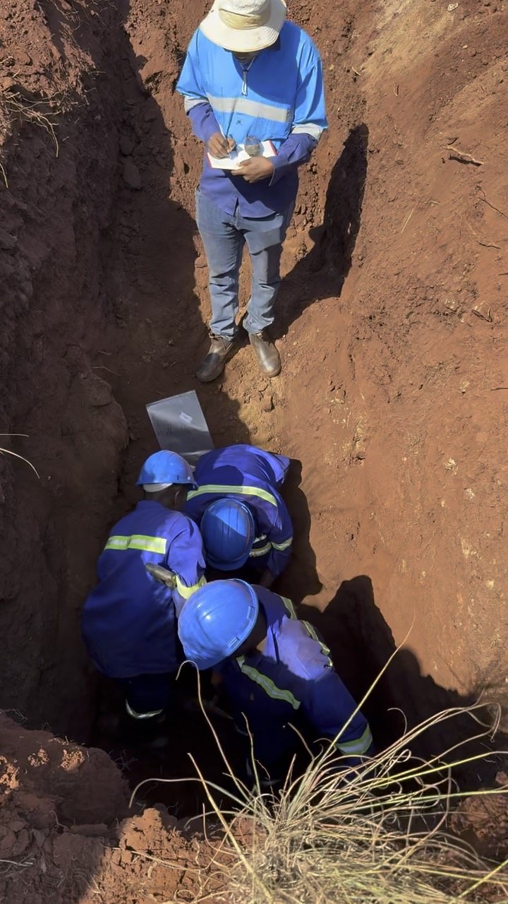 A geologist logging notes beside a pit while workers take samples at the bottom