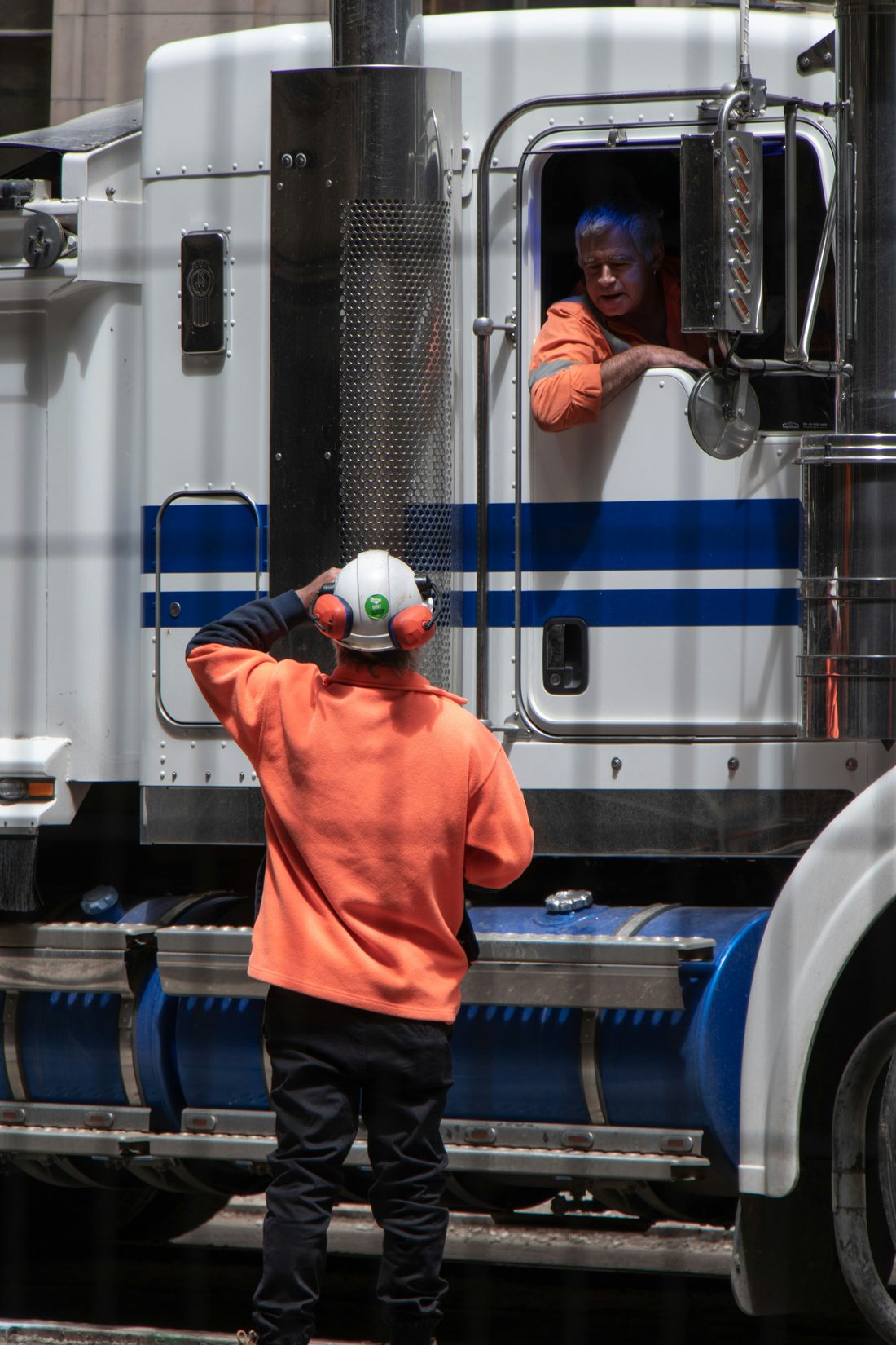 The driver of a white long-haul truck leans out of the cab window to speak with a site worker in a hard hat and high-visibility jacket
